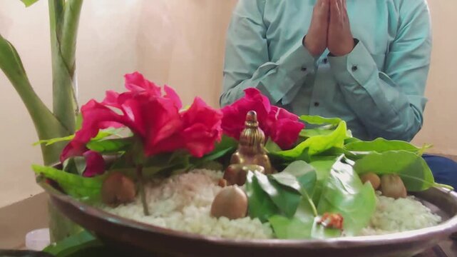 Devotee in a formal shirt sitting in a namaskar prayer posture behind a brass Balkrushna idol adorned with red hibiscus and sacred bael leaves during a home pooja.
