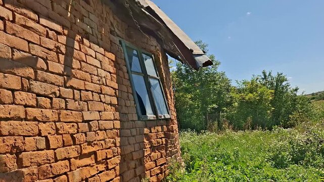 Old brick house exterior with weathered masonry, a damaged roof edge, blue framed window, and overgrown green vegetation surrounding the abandoned rural property