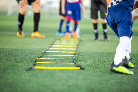 Selective focus of a yellow speed agility ladder placed on green artificial turf field for footwork training drills with blurred kids practicing soccer in background.