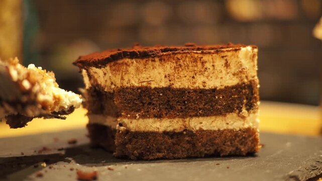 Taking a bite of tiramisu cake with metal fork on black stone board in restaurant setting.