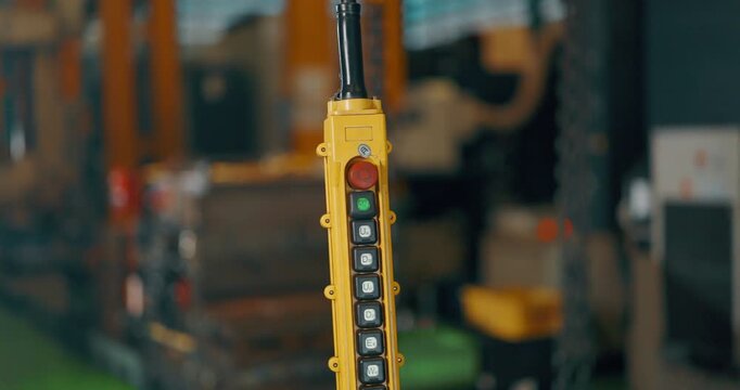 Close up of the yellow pendant control station of an overhead travelling crane hanging from its cable, with push buttons and a red emergency stop, inside an industrial workshop.

