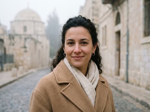 Middle Eastern woman with curly hair wearing warm autumn coat smiling on old historic street in Jerusalem, capturing authentic culture, city exploration, and travel vibes