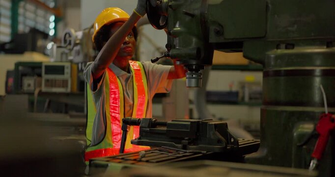 A Black female machinist in a hi vis vest, hard hat and safety glasses sets a metal workpiece in the machine vice of an industrial drilling machine and operates the spindle in a factory.
