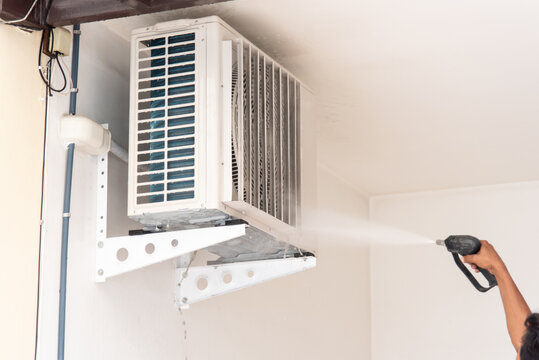 Technician washing an outdoor air conditioner condenser unit high pressure water during maintenance service to remove dirt improve cooling efficiency