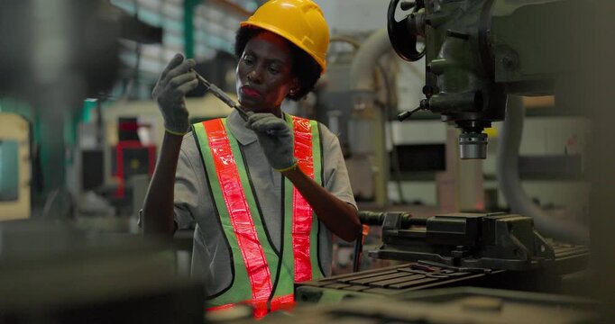 A Black female machinist in a hi vis vest and hard hat holds a drill bit up to inspect its cutting edge and reaches to the chuck of an industrial drilling machine in a factory workshop.
