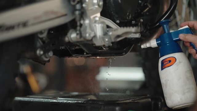 Mechanic sprays degreaser from a pressure bottle onto the engine and swingarm of a motorcycle during a workshop clean.