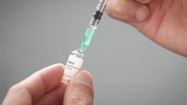 Fingers hold a small glass ampoule of testosterone while a syringe needle draws the oil out of it