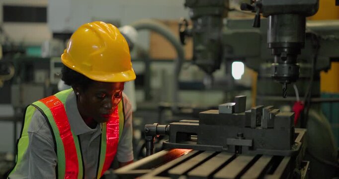 A Black female machinist in a hi vis vest, hard hat and safety glasses sets a metal workpiece in the machine vice of an industrial drilling machine and operates the spindle in a factory.
