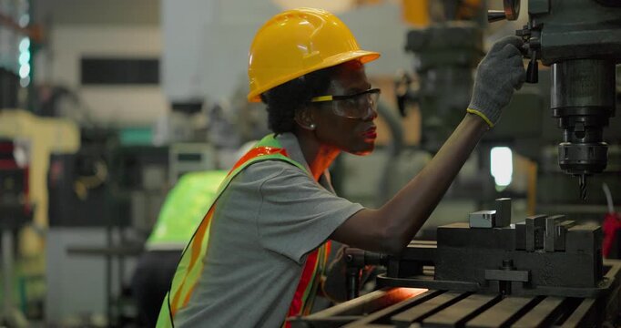 A Black female machinist in a hi vis vest and hard hat holds a drill bit up to inspect its cutting edge and reaches to the chuck of an industrial drilling machine in a factory workshop.
