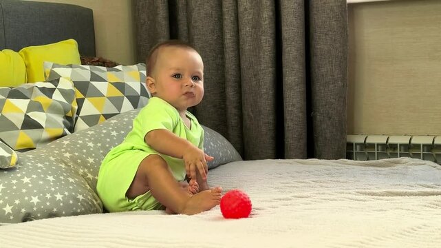 Curious baby sits on a bed with a red sensory ball at home, infant development, motor skills, playful learning and childhood exploration.