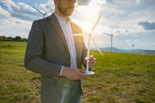 A Caucasian man in a suit jacket and glasses holds a small model of a wind turbine while standing in a grassy field with large wind turbines visible on the horizon
