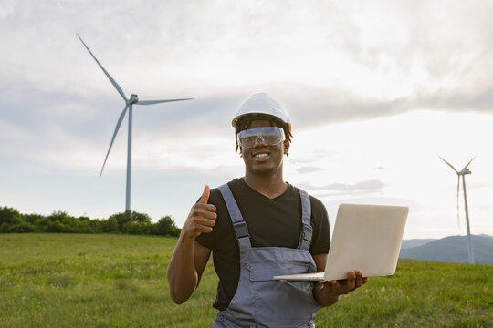A smiling African American man in work clothes and safety gear holds a laptop and gives a thumbs-up gesture in a field with wind turbines