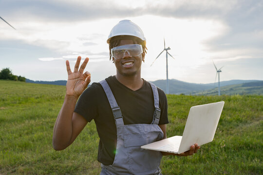 A smiling African American man in safety gear holds a laptop and gestures an ok sign in a field with wind turbines