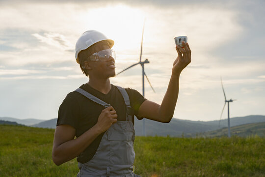 A man in a hard hat and overalls examines a lightbulb in a field with wind turbines generating clean energy