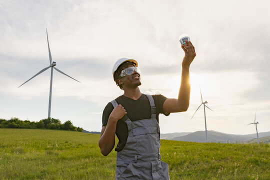 A smiling African American man in work clothes and safety gear holds up an LED light bulb while standing in a field with wind turbines