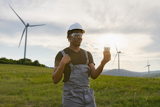 A man in work overalls and safety gear inspects an LED light bulb with wind turbines in the background