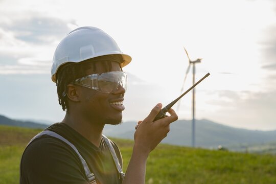 A smiling African American man wearing a hard hat and safety glasses speaks into a radio with wind turbines in the background