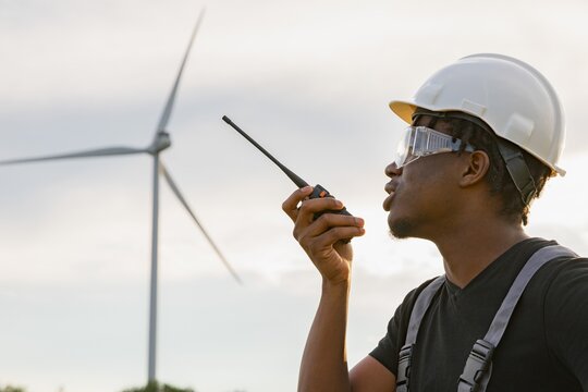 A male technician wearing protective gear communicates via a walkie-talkie while working at a wind farm facility