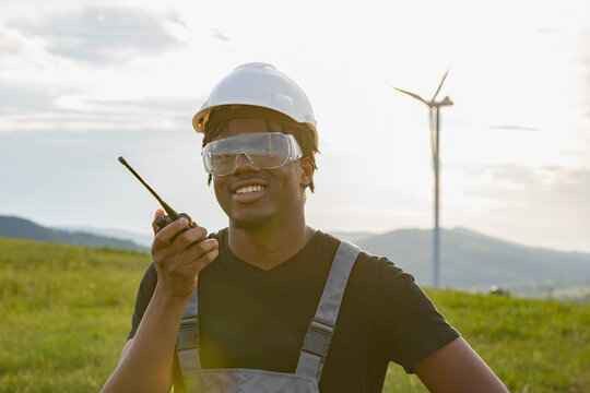 A smiling African American man wearing a hard hat and safety goggles holds a radio while standing in a field with a wind turbine in the background