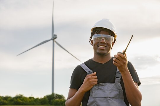 A smiling African American man in a hard hat and safety glasses holds a radio while standing in front of a wind turbine