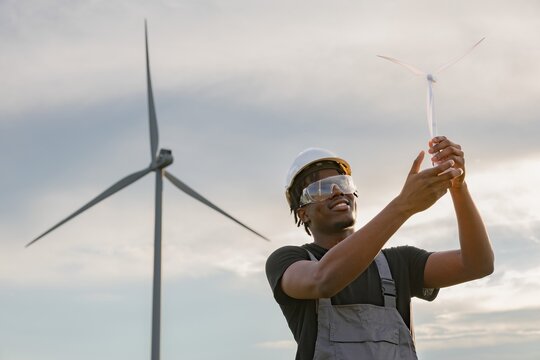 A smiling African American man in safety gear holds a miniature wind turbine, contemplating its design with a full-scale turbine in the background