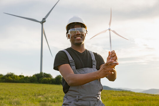 A smiling African American man in work clothes and safety gear holds a small wind turbine model in a field with large windmills behind him
