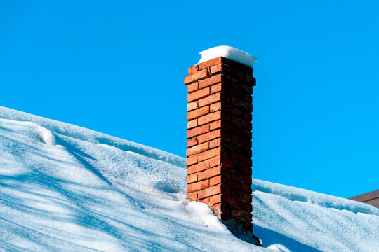 Architectural winter detail of a tall red brick chimney rising from a thick snow-covered sloped roof, topped with a perfect white snow cap and small icicle drip