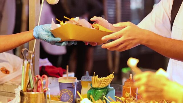 Street food vendor adding sauce to tasty snacks on skewers and serving customer.