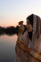 Side profile portrait of a beautiful young Indian woman in traditional pink ethnic dress and embroidered dupatta, holding an orange marigold flower by the lake during sunset for Diwali festival celebr © Alya
