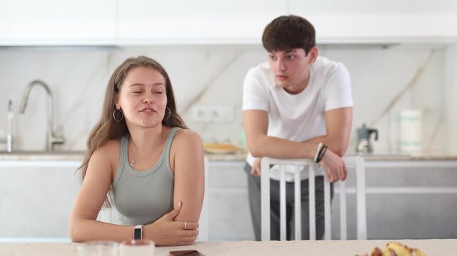 Sad young girl sitting at the table while her boyfriend is quarreling with her standing behind in the kitchen 