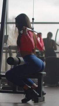 Woman seated on cable pulldown machine gripping wide bar overhead, showing defined shoulders and upper back in modern fitness studio lit by backlit window