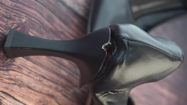 Damaged high heel shoe showing broken heel and torn leather on dark wooden floor background.
