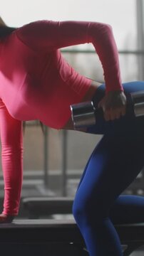 Woman performing bent-over one-arm dumbbell row on bench in gym with bright pink top and blue leggings, low eye-level vertical framing emphasizing torso arm and dumbbell