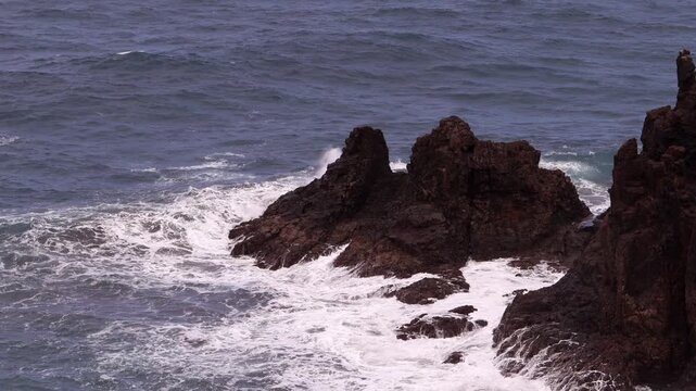 Sharp dark rocks jut out of the open sea, waves washing gently around their base under a calm sky. Jagged Rocks Standing in Open Ocean Water