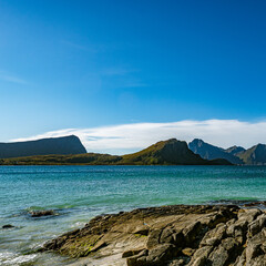 Panoramic view of a rocky coastline with turquoise sea water and majestic mountain peaks under a clear blue sky in Norway with copy space © Quality by Simon