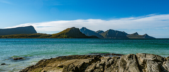 Panoramic view of a rocky coastline with turquoise sea water and majestic mountain peaks under a clear blue sky in Norway with copy space © Quality by Simon