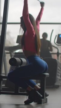 Focused woman performing seated resistance exercise in modern fitness studio, stretching arms overhead and engaging lower body muscles during strength training session