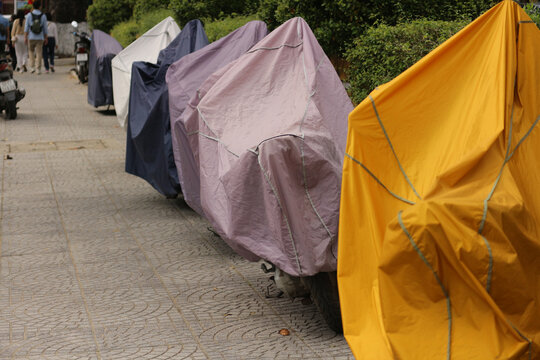 Row of parked motorbikes and scooters covered with colorful waterproof tarpaulin covers on a city street pavement in Hoi An, Vietnam.