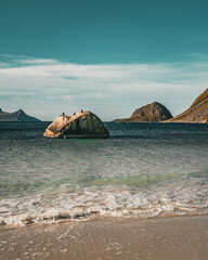 Vertical view of a scenic ocean beach with a solitary rock in the sea and mountain peaks under a calm blue sky in Norway with copy space © Quality by Simon