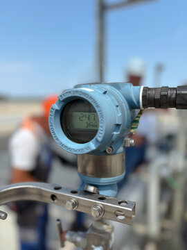 A close-up photograph showing a digital pressure transmitter gauge mounted on metallic piping, with a clear LCD screen displaying pressure readings in bar. Selective focus
