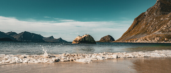 Panoramic view of gentle ocean wave splashing on sandy beach with solitary rock and majestic mountains under blue sky in Norway © Quality by Simon
