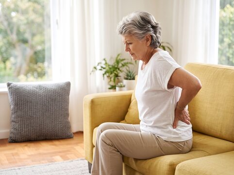 Professional Stock Image of an Elderly Woman Sitting on a Yellow Sofa While Holding Her Lower Back and Suffering From Chronic Waist Pain, Muscle Strain, and Physical Fatigue