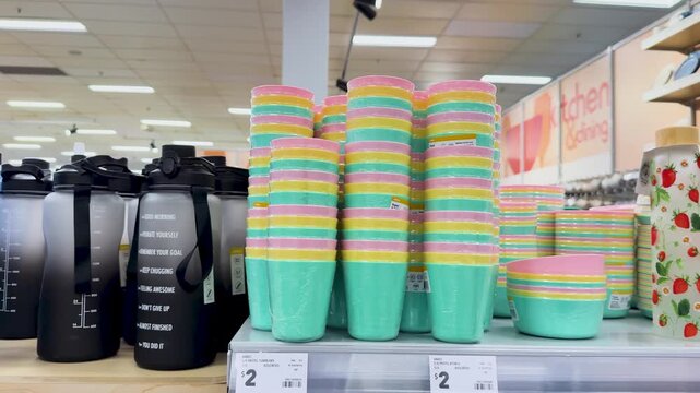 Colorful plastic cups stacked neatly on store shelf, kitchenware and water bottles in background.