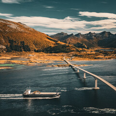 Aerial drone view of a cargo vessel sailing under a long concrete bridge across a sea fjord in scenic Norway landscape with copy space © Quality by Simon