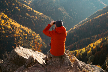 Fototapeta premium Man sitting on a rocky ledge and looking through binoculars at an autumn mountain landscape.