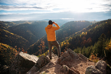 Fototapeta premium Man standing on a rocky ledge and looking through binoculars at an autumn mountain landscape.