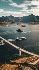 Vertical drone view of a red car driving on a long bridge over a fjord while a cargo ship passes underneath in scenic Norway landscape with copy space © Quality by Simon