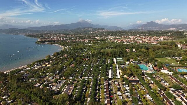 Rows of caravans fill dense campground beside curved turquoise swimming pool, sandy beach lines lake shore near distant town, hazy mountains rise beyond Lake Garda, Italy. Aerial drone wide shot