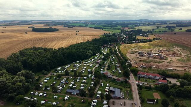 Rows of white caravans fill forested campground beside golden wheat fields, construction site with heavy machinery borders quiet village road near Baltic Sea, Germany. Aerial drone backwards shot