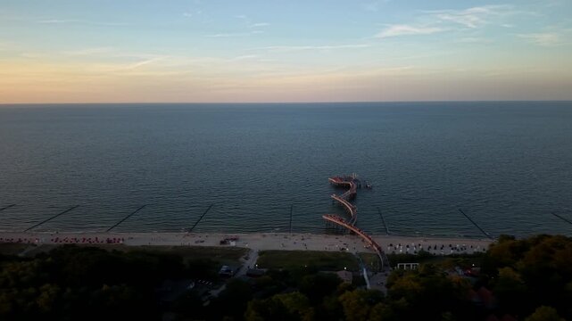 Curved wooden pier winds across calm evening sea, rows of striped beach chairs line long sandy shoreline beneath fading orange sunset sky above Usedom coastline, Germany.Aerial drone lateral wide shot